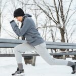 Person jogging on a snowy path during winter sunrise, dressed in warm workout clothes.