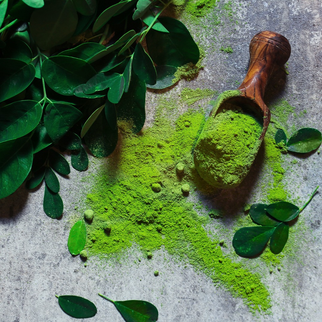 Fresh moringa leaves and a spoonful of moringa powder displayed on a wooden surface.